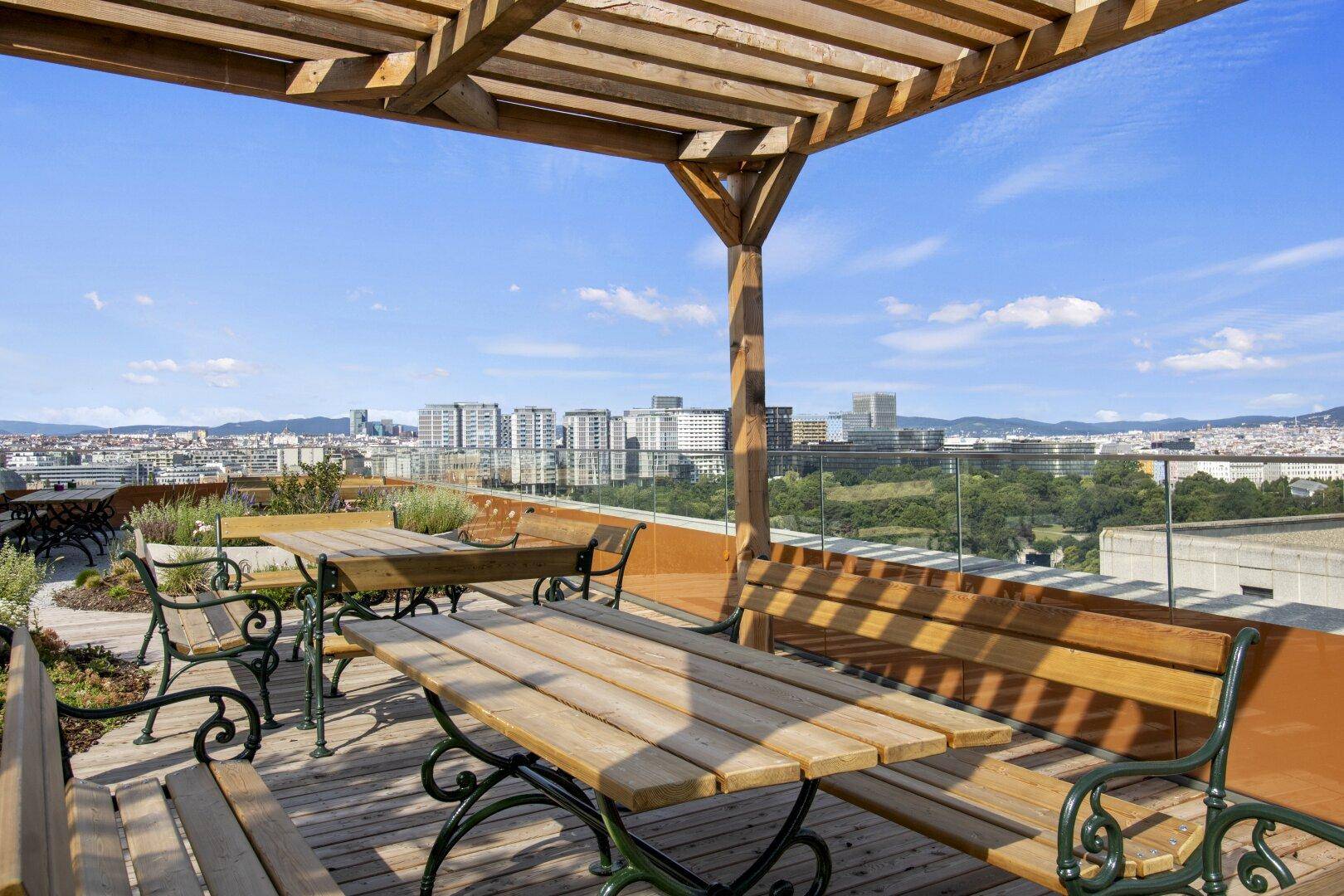 Gemütliche Dachterrasse mit Holzpergola, Picknicktischen und weitem Blick über die Stadt und grüne Hügel.