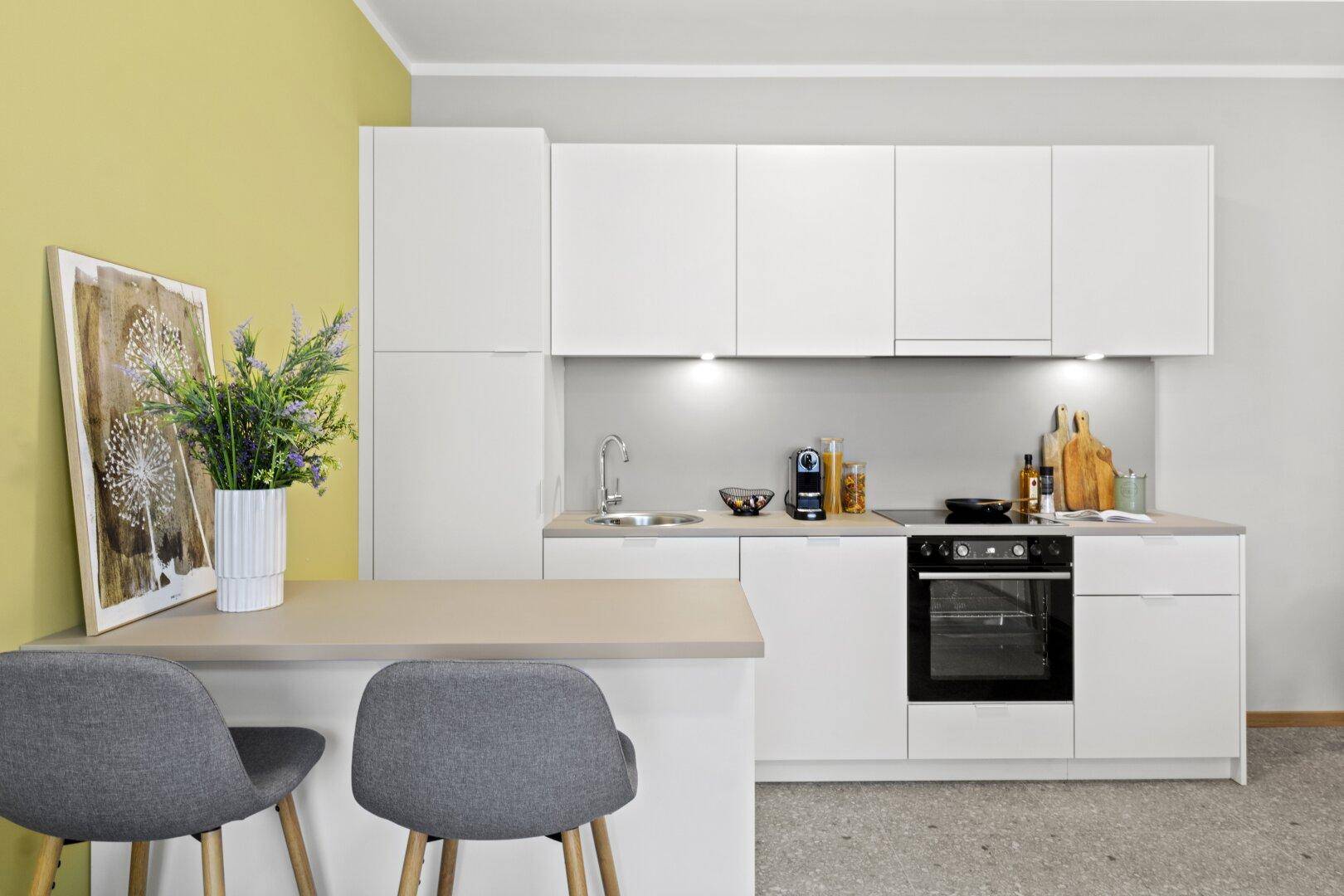 Moderne, helle Küche mit weißen Schränken, gelber Akzentwand und integriertem Essbereich.