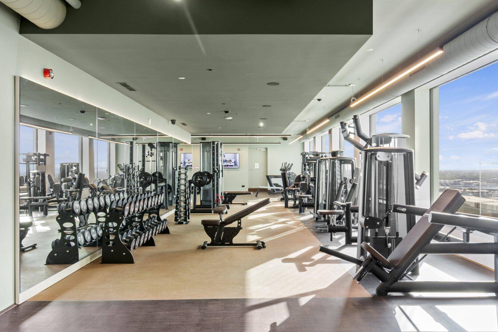 Geräumiger Fitnessraum mit modernen Trainingsgeräten, großen Fenstern und Spiegelflächen.