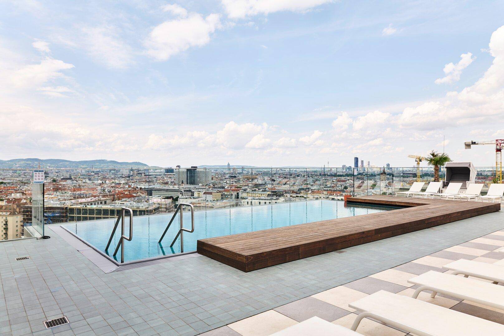 Weitläufiger Dachpoolbereich mit Liegestühlen, Holzdeck und Panoramablick über die Stadtlandschaft.