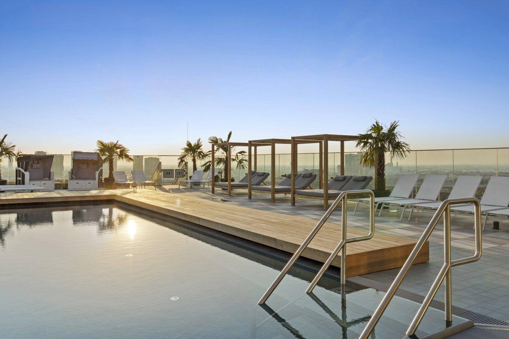 Dachterrassenpool mit Liegestühlen und Palmen, bietet einen weiten Blick über die Stadt bei Sonnenuntergang.