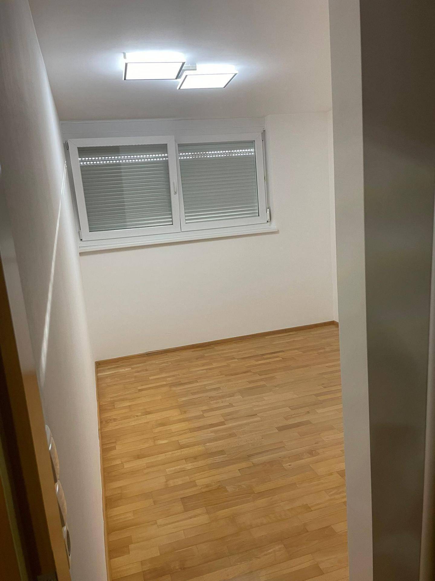 Leeres Zimmer mit Holzparkettboden und Fenstern, die mit Jalousien versehen sind.