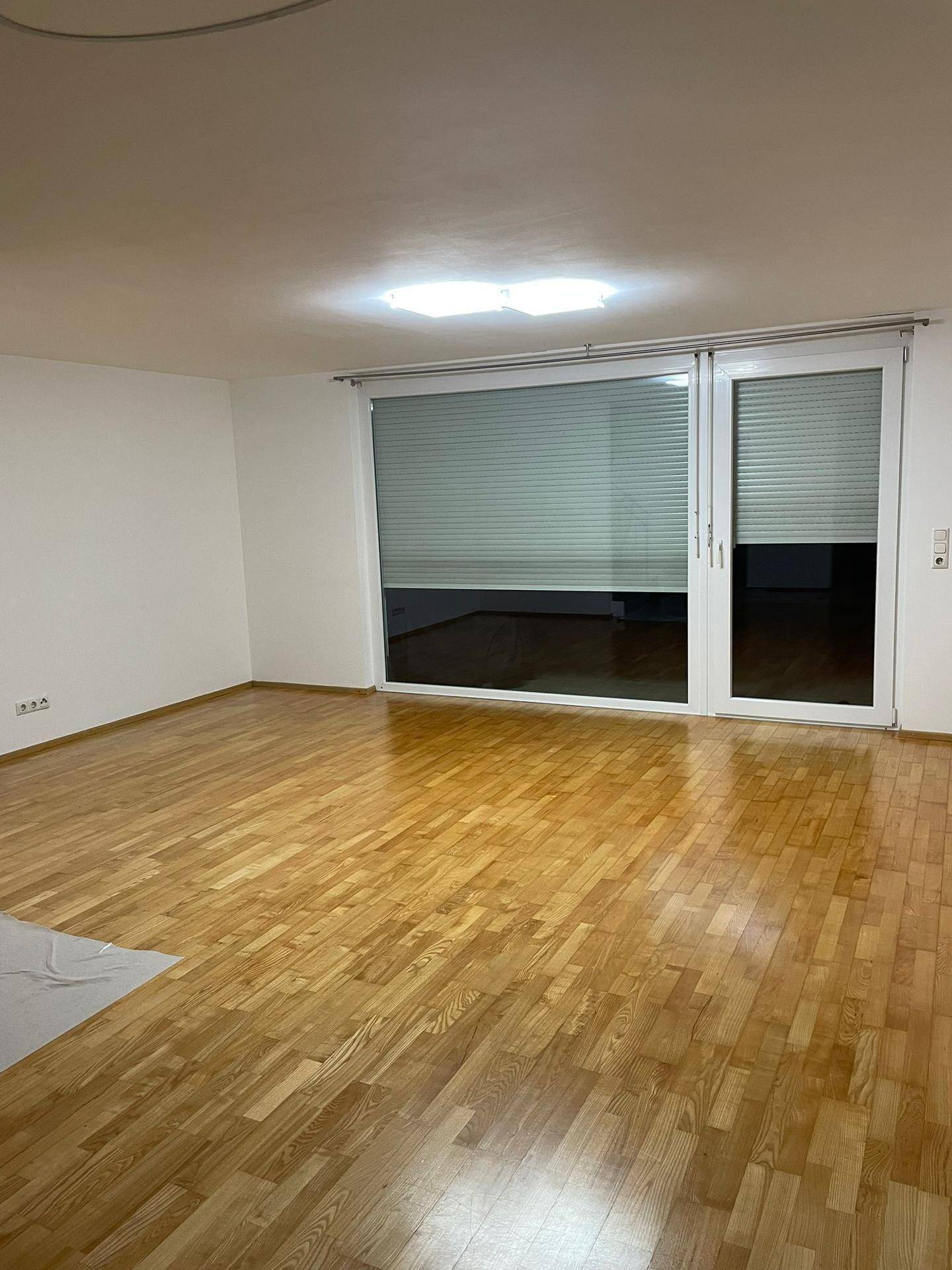 Helles, leeres Wohnzimmer mit hellem Holzparkett und großen Fenstern mit Jalousien.