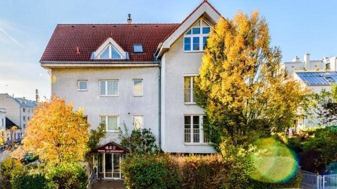 Vorderansicht eines modernen Gebäudes mit rotem Ziegeldach und großen Fenstern, umgeben von Herbstbäumen.