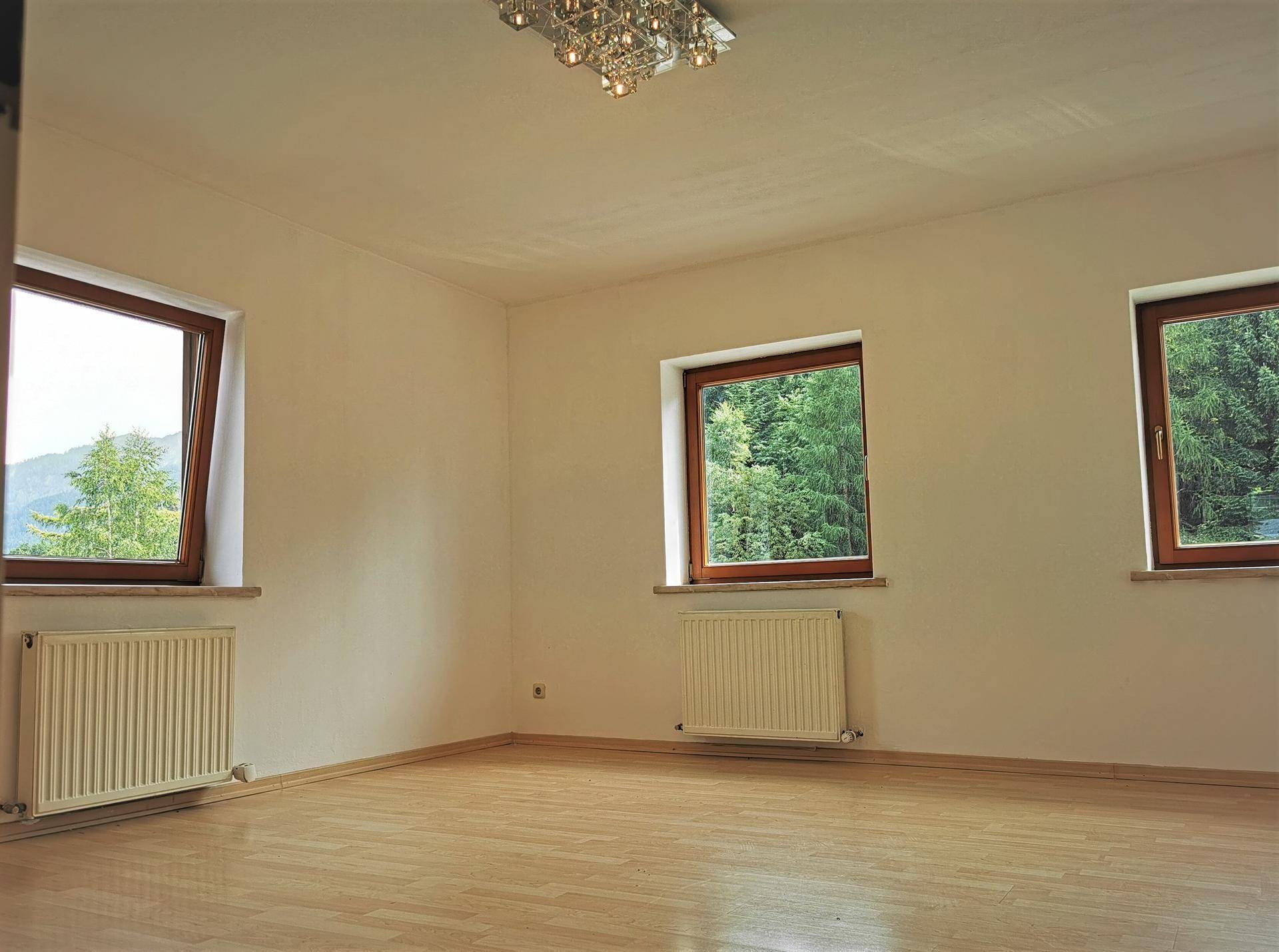 Geräumiger, heller Raum mit Parkettboden und drei großen Fenstern mit Blick ins Grüne.