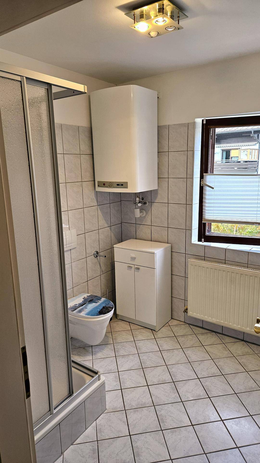 Badezimmer mit grauen Fliesen, Duschkabine, WC und einem Fenster für natürliche Belüftung und Licht.