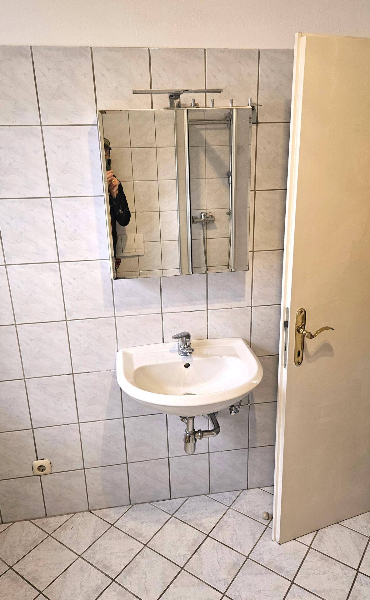 Badezimmer mit Wandwaschbecken, Spiegelschrank und hellen Fliesen an den Wänden, praktisch eingerichtet.