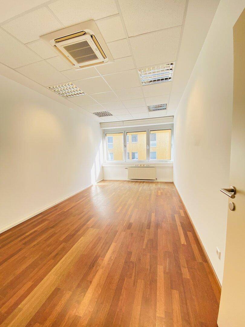 Kleineres Büro mit Holzboden, großen Fenstern und Klimaanlage, ideal für Einzelarbeitsplätze oder Besprechungen.