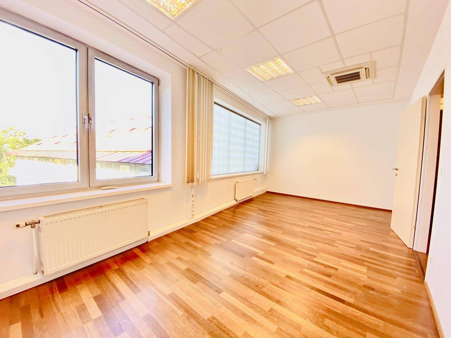 Helles Büro mit Holzboden, großen Fenstern und Heizkörpern, bietet viel Tageslicht für eine angenehme Arbeitsumgebung.