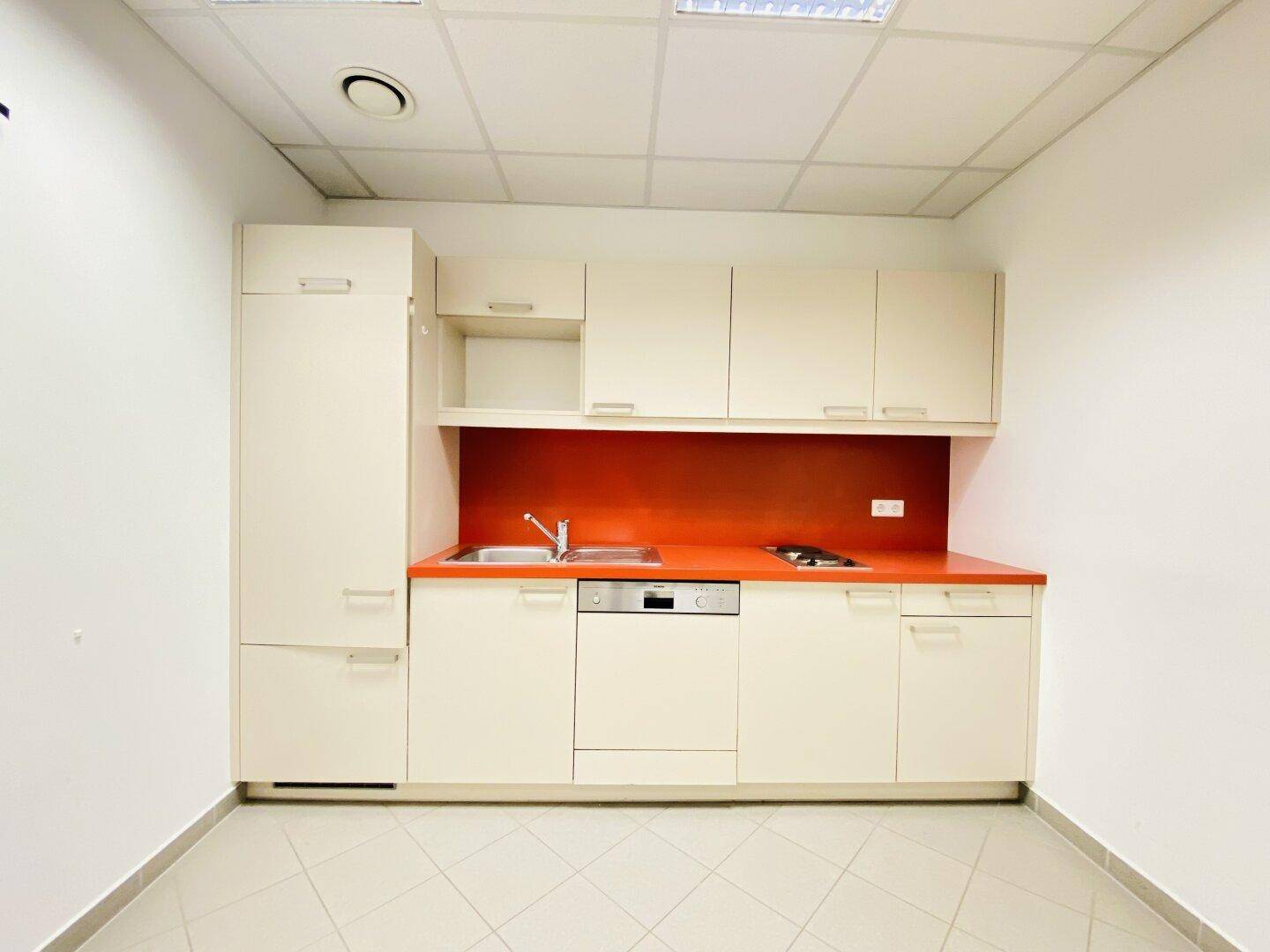Funktionale Küchenzeile mit beigen Schränken, roter Rückwand und integriertem Geschirrspüler, ideal für Pausenräume.