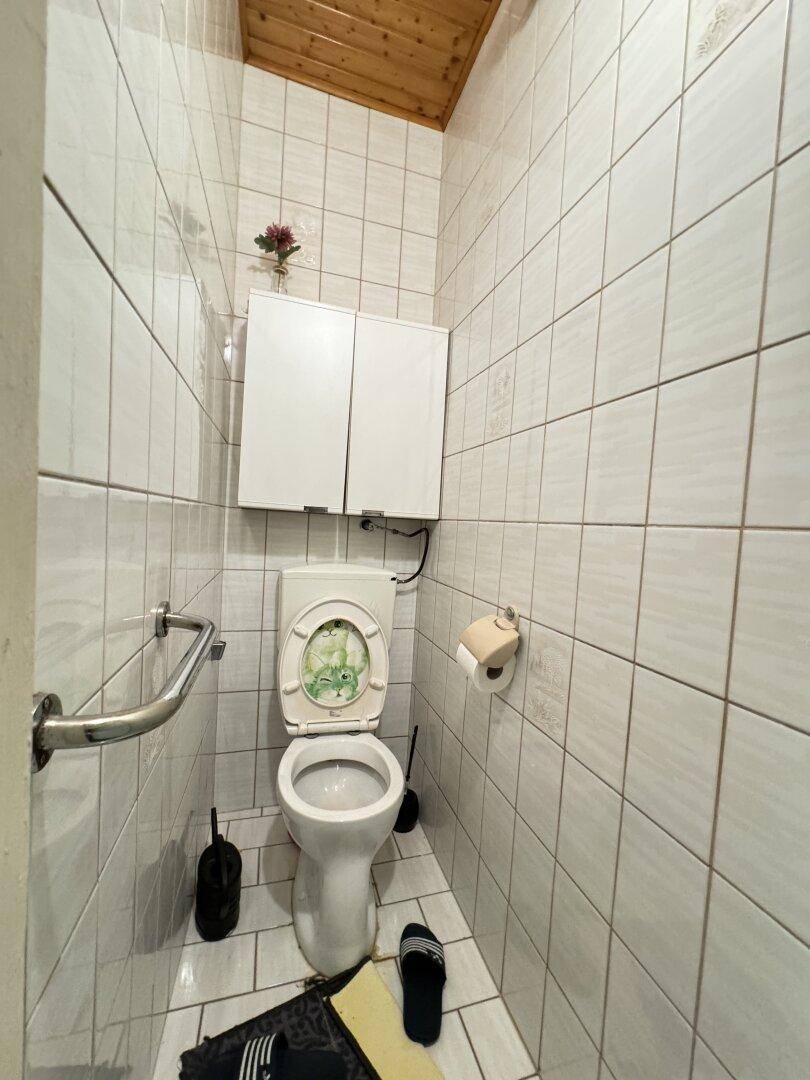 Separates WC mit Toilette, weißen Fliesen und einer kleinen Ablage über dem Spülkasten.