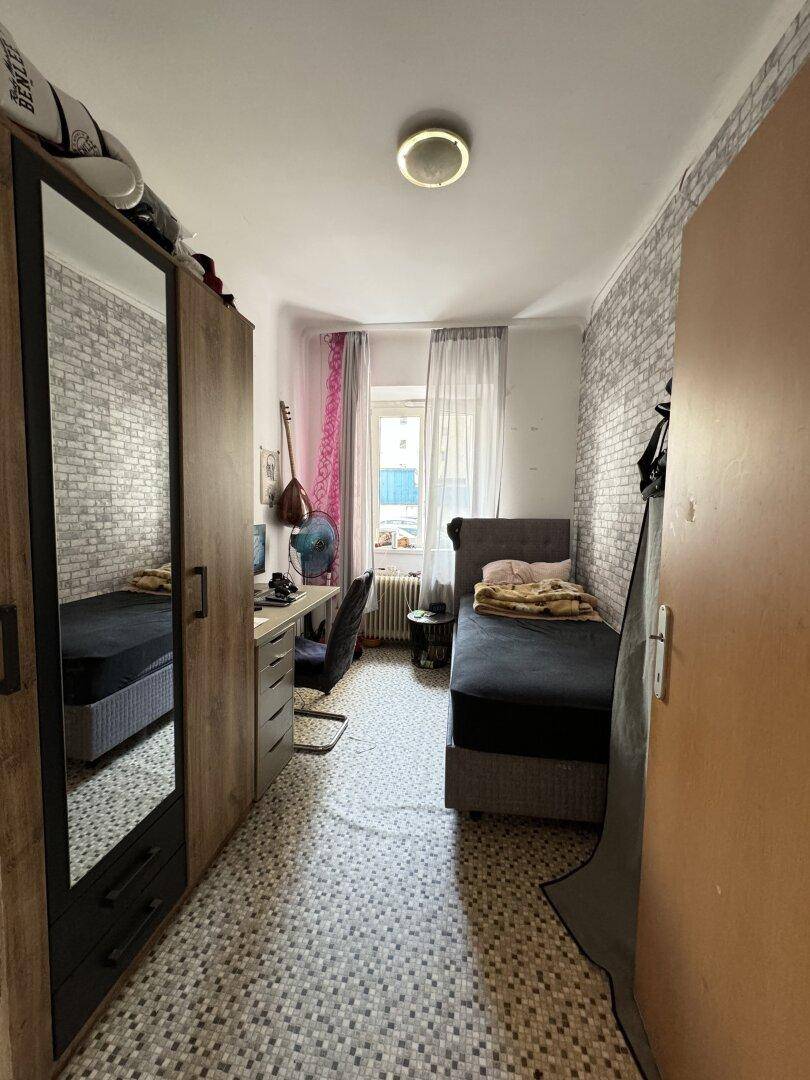 Schlafzimmer mit Einzelbett, Schreibtisch und Kleiderschrank, mit gemustertem Bodenbelag.
