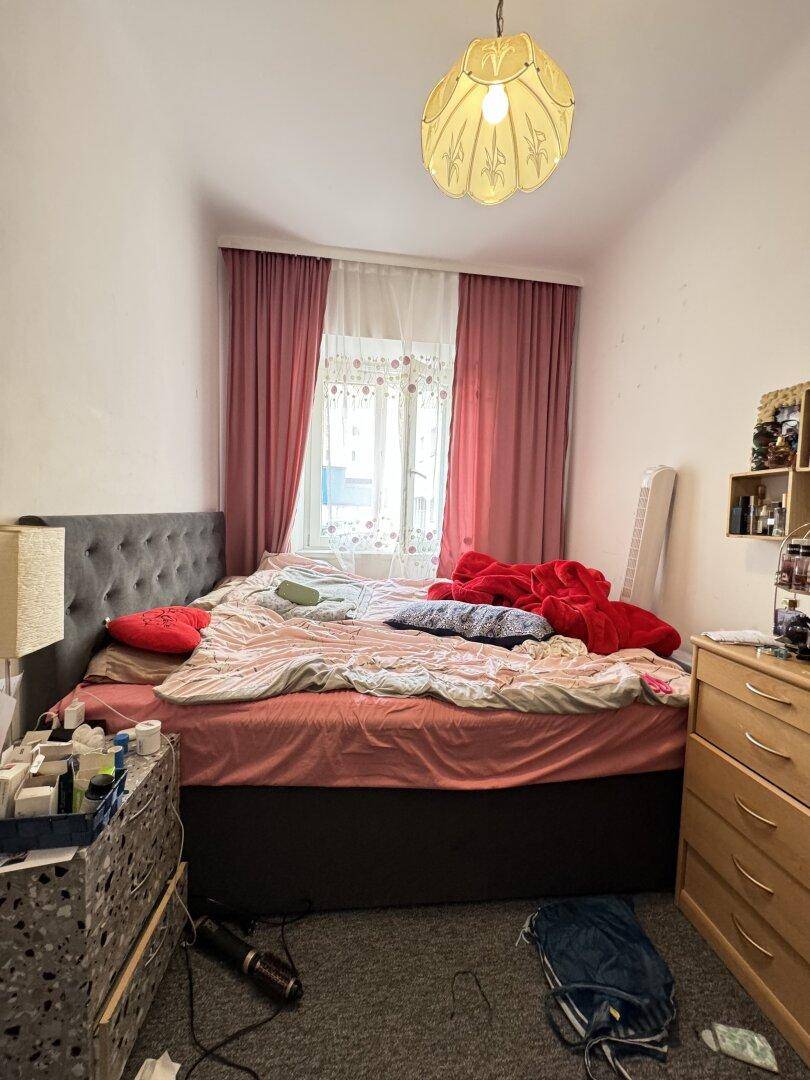 Gemütliches Schlafzimmer mit Doppelbett, Kommode und rosa Vorhängen, beleuchtet durch ein Fenster.