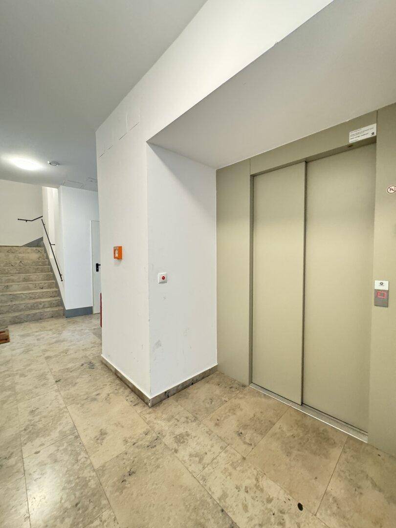 Eingangsbereich mit geschlossener Aufzugstür und Treppenhaus, hell gefliest.