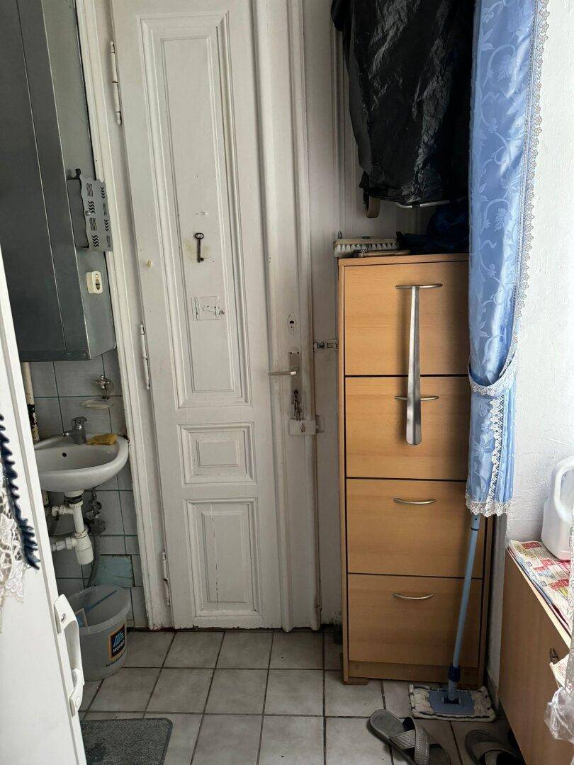 Eingangsbereich mit alter Holztür, kleinem Waschbecken und Fliesenboden.