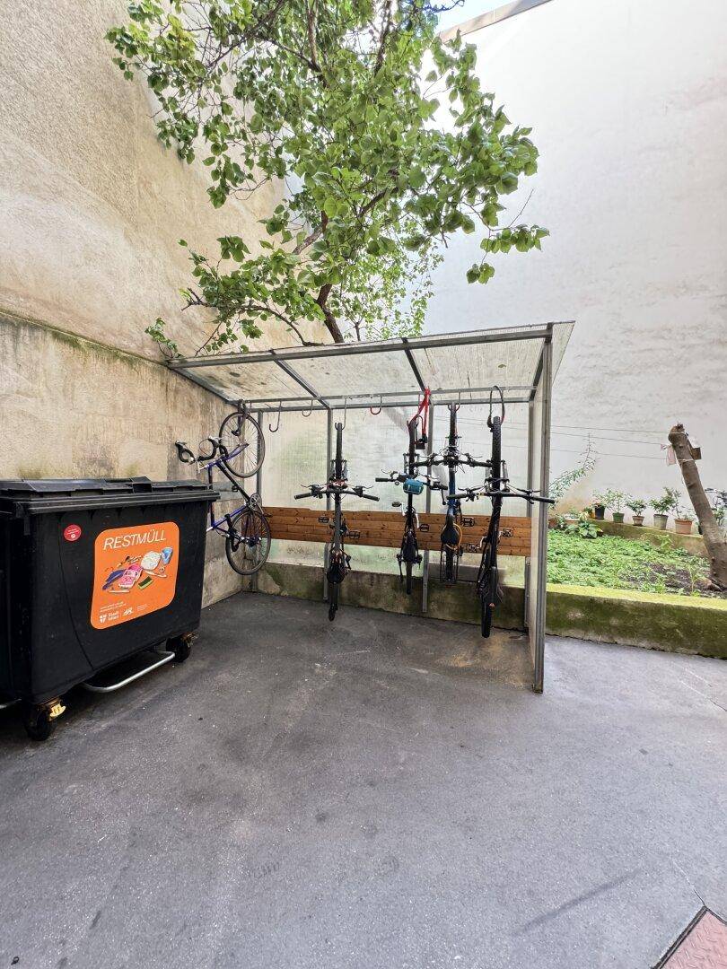 Außenbereich mit überdachtem Fahrradabstellplatz und Mülltonnen, umgeben von einer Mauer und etwas Grün.