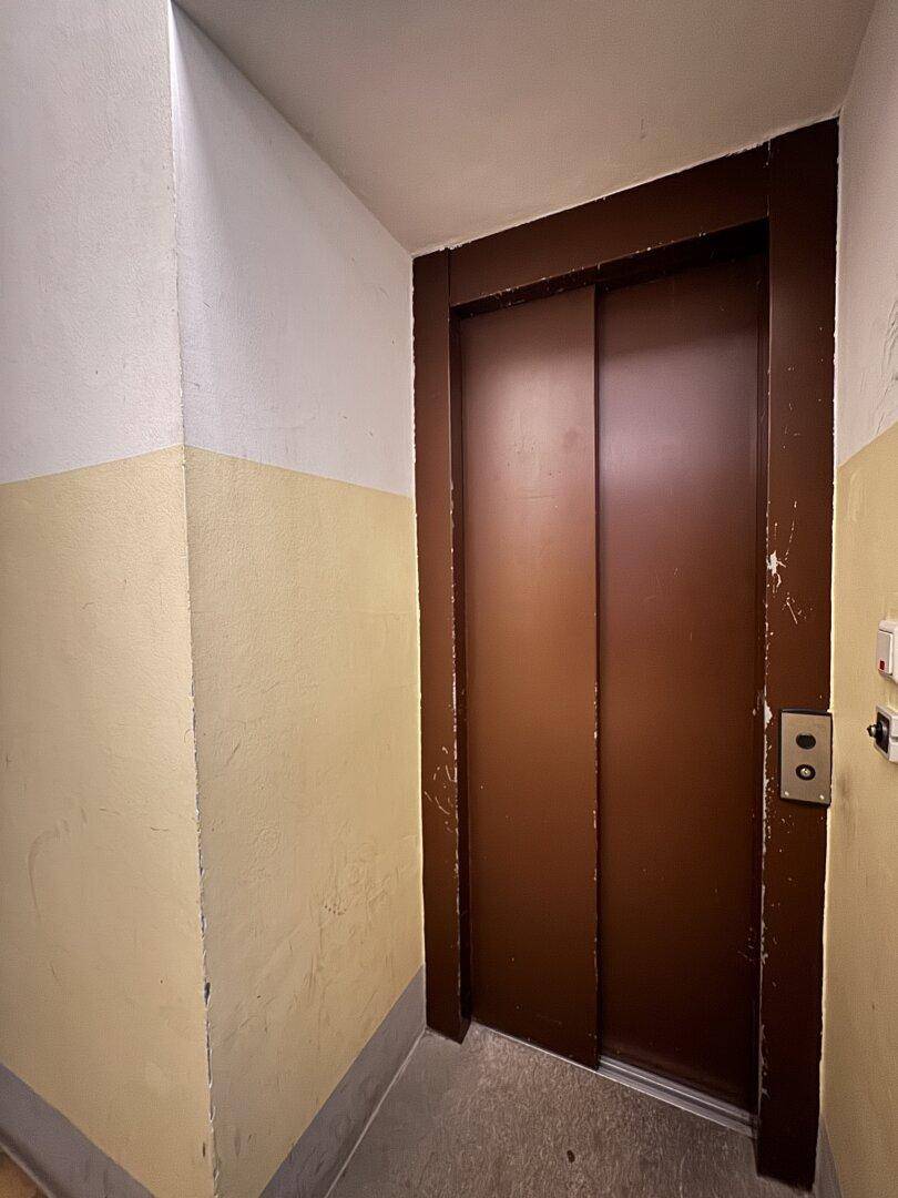 Aufzug im Treppenhaus eines Altbaus mit braunen Türen und gelb gestrichenen Wänden.