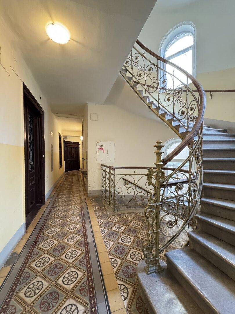 Elegantes Treppenhaus eines Altbaus mit geschwungenem Geländer und gemusterten Fliesen im Eingangsbereich.