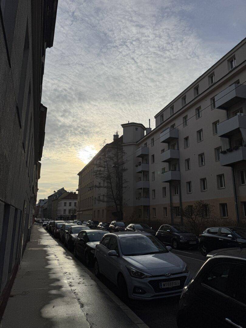 Sonnenuntergang über einer belebten Stadtstraße mit parkenden Autos und hohen Wohngebäuden.