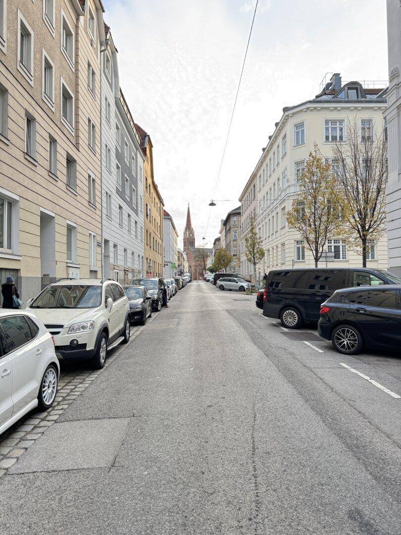 Eine belebte Stadtstraße mit parkenden Autos und hohen Gebäuden, die zu einem Kirchturm führt.