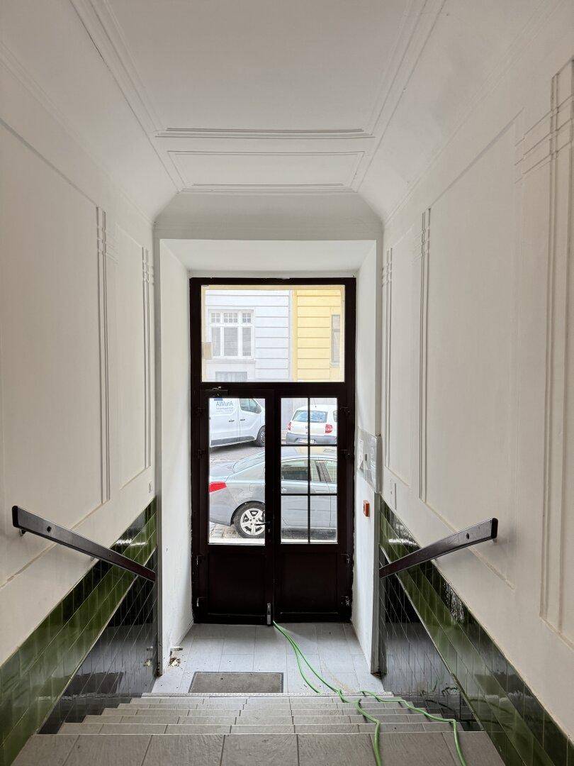 Eingangsbereich mit Treppe, grünen Fliesen an den Wänden und einer Holztür zum Außenbereich.