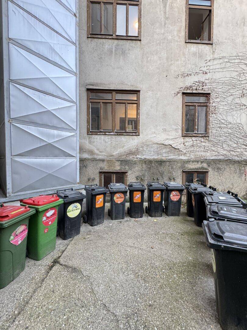 Hinterhof eines Gebäudes mit mehreren Mülltonnen und einer schlichten Fassade mit Fenstern.