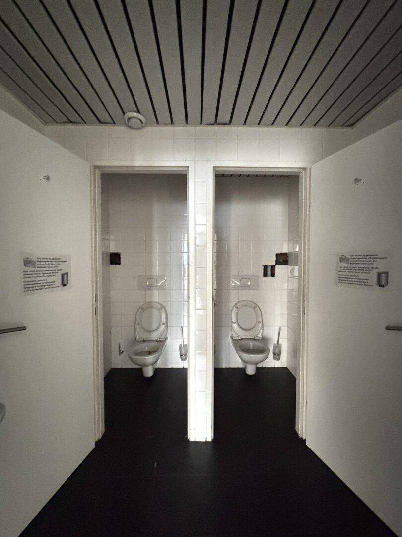 Zwei separate Toilettenkabinen mit weißen Fliesen und dunklem Boden in einem Sanitärbereich.