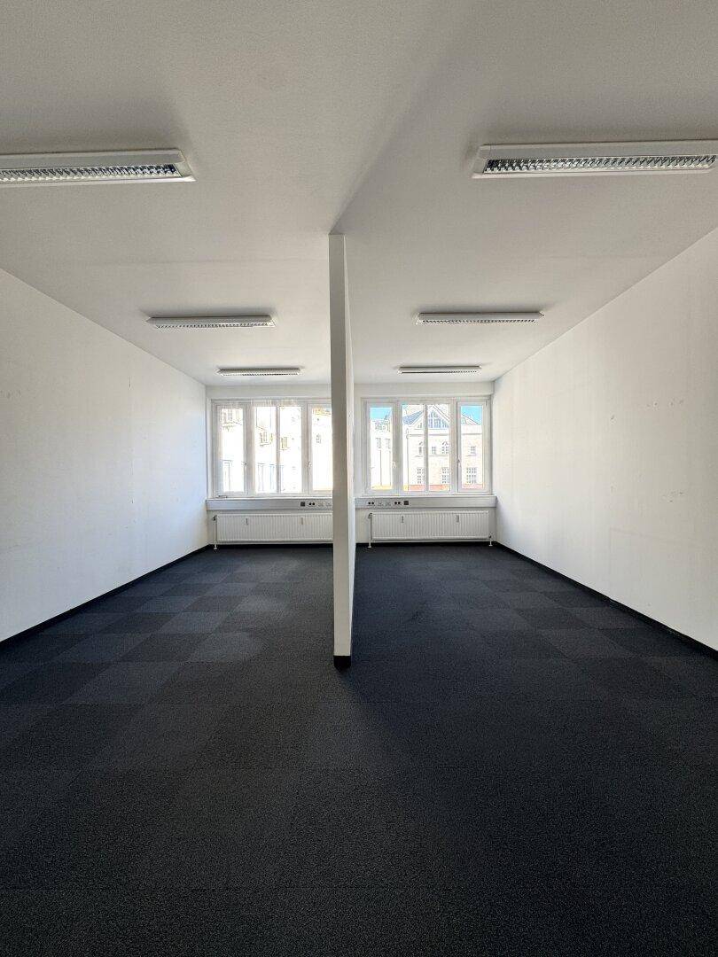 Helles Büro mit Teppichfliesen, großen Fenstern und einer zentralen Säule, die den Raum teilt.