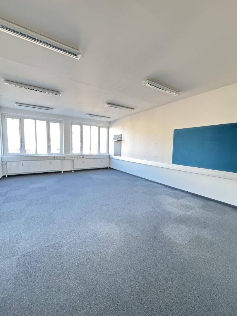 Geräumiges, leeres Büro mit Teppichfliesen, großen Fenstern und einer blauen Wandtafel.