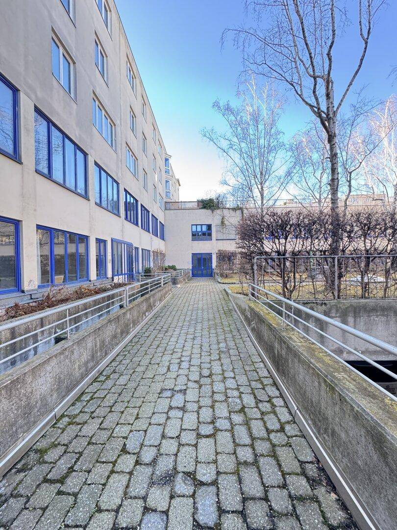 Gepflasterter Weg führt entlang der Gebäudefassade mit blauen Fenstern und Geländern.