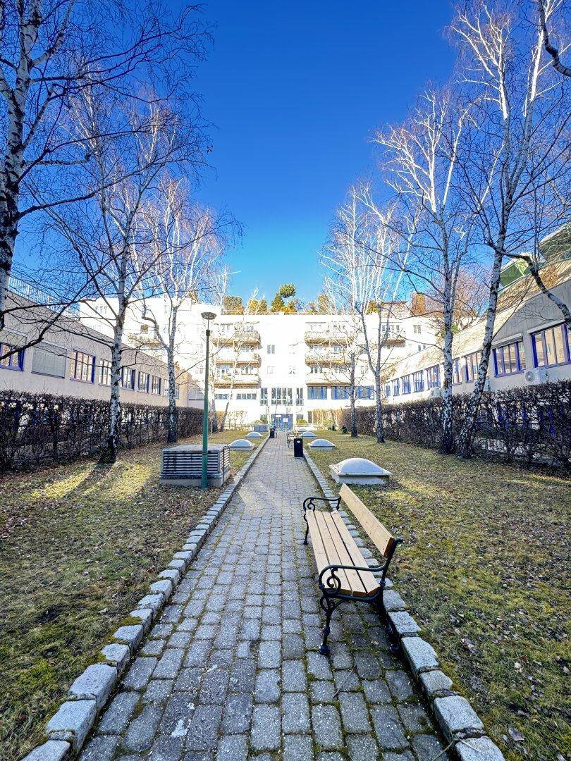 Innenhof mit gepflastertem Weg, Bäumen, einer Bank und dem Gebäudekomplex im Hintergrund.