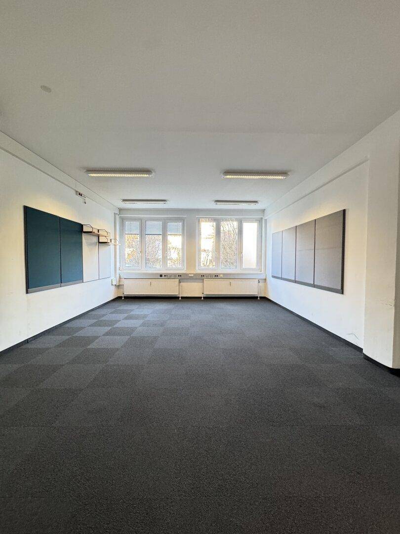 Geräumiges, leeres Büro mit Teppichfliesen, großen Fenstern und Wandtafeln.
