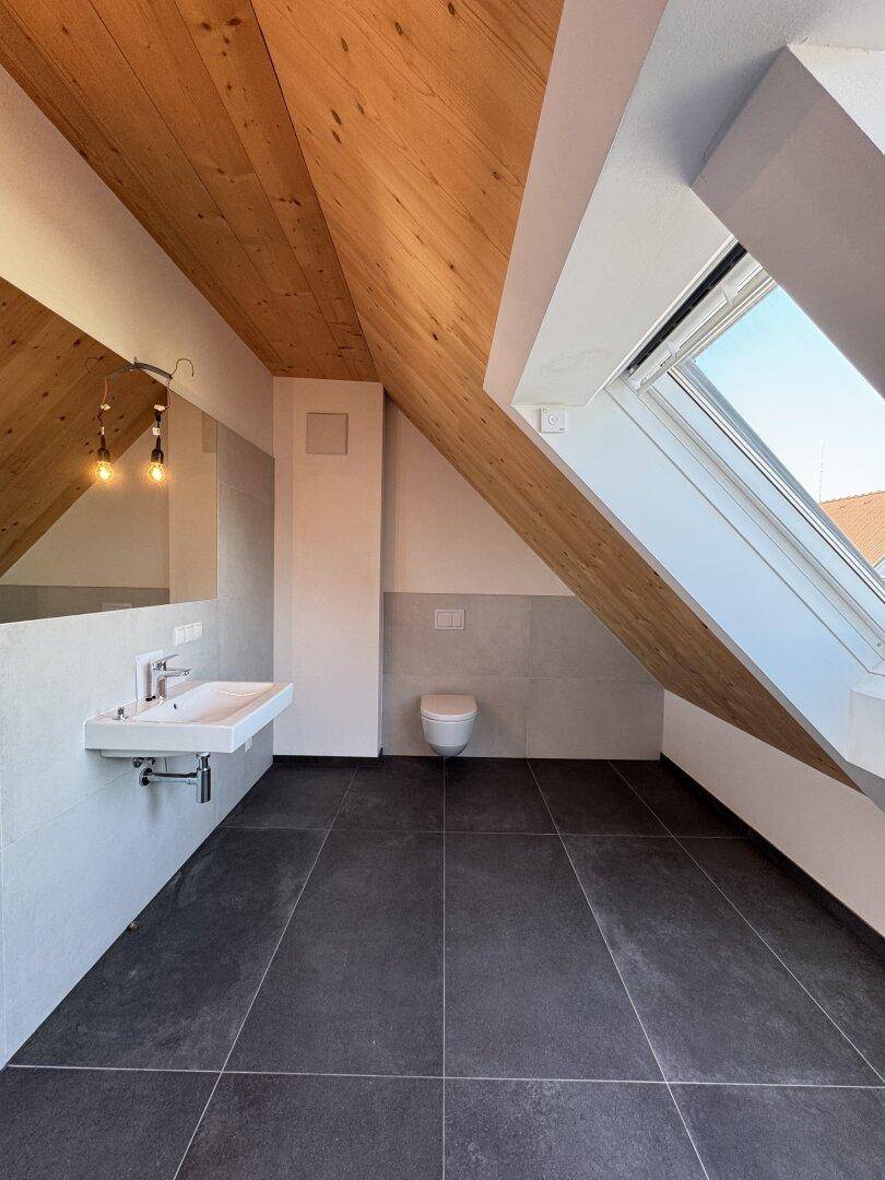 Modernes Badezimmer mit dunklen Fliesen, Holzbalkendecke, wandhängendem WC und großem Dachfenster.
