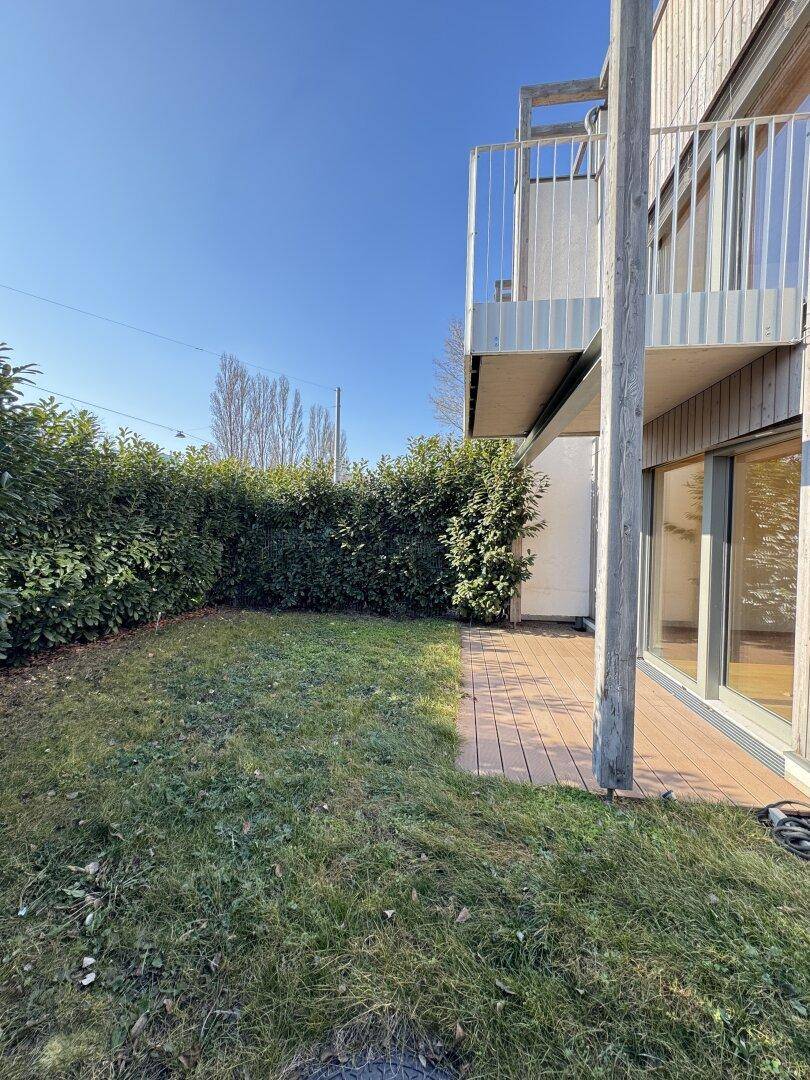 Kleiner Gartenbereich mit Holzterrasse und Hecke neben dem Gebäude, darüber ein Balkon.