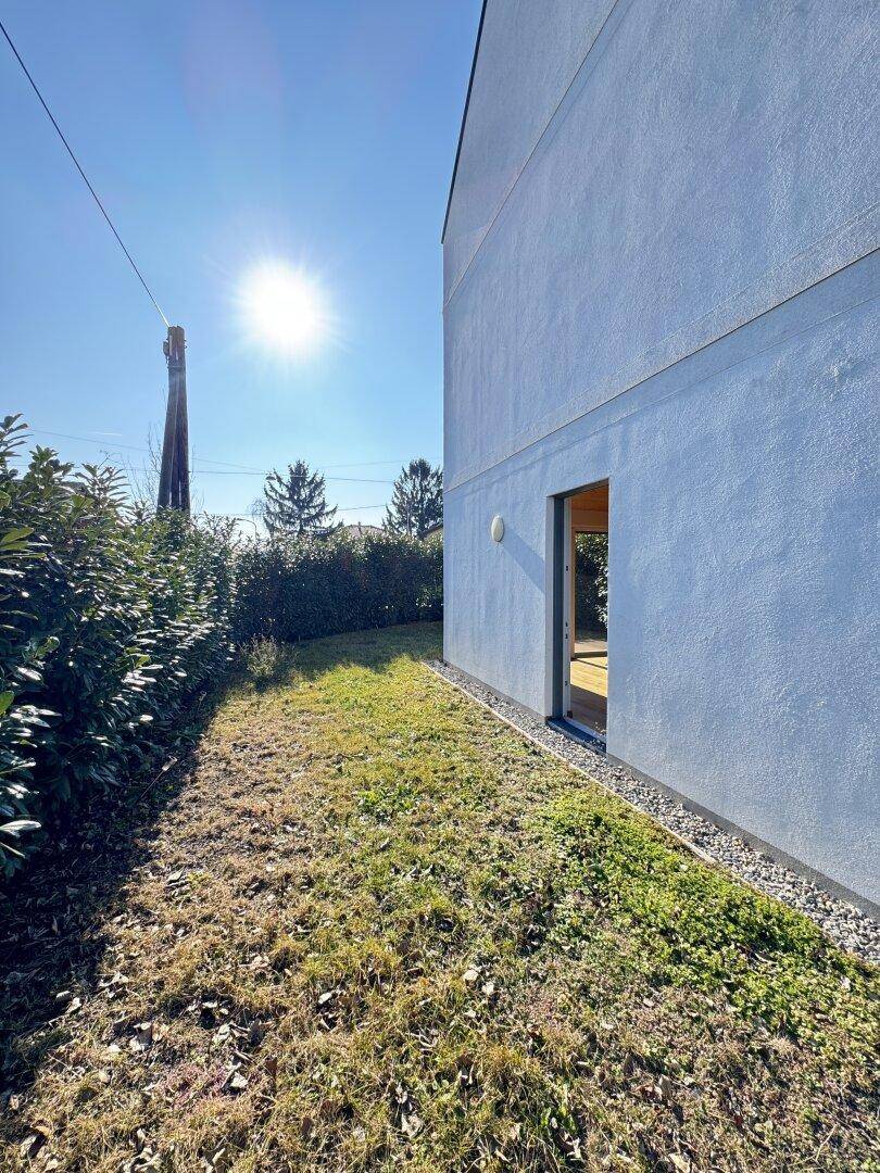 Moderne Gebäudeseite mit weißer Fassade, umgeben von einer Hecke und einem sonnigen Rasenstück.