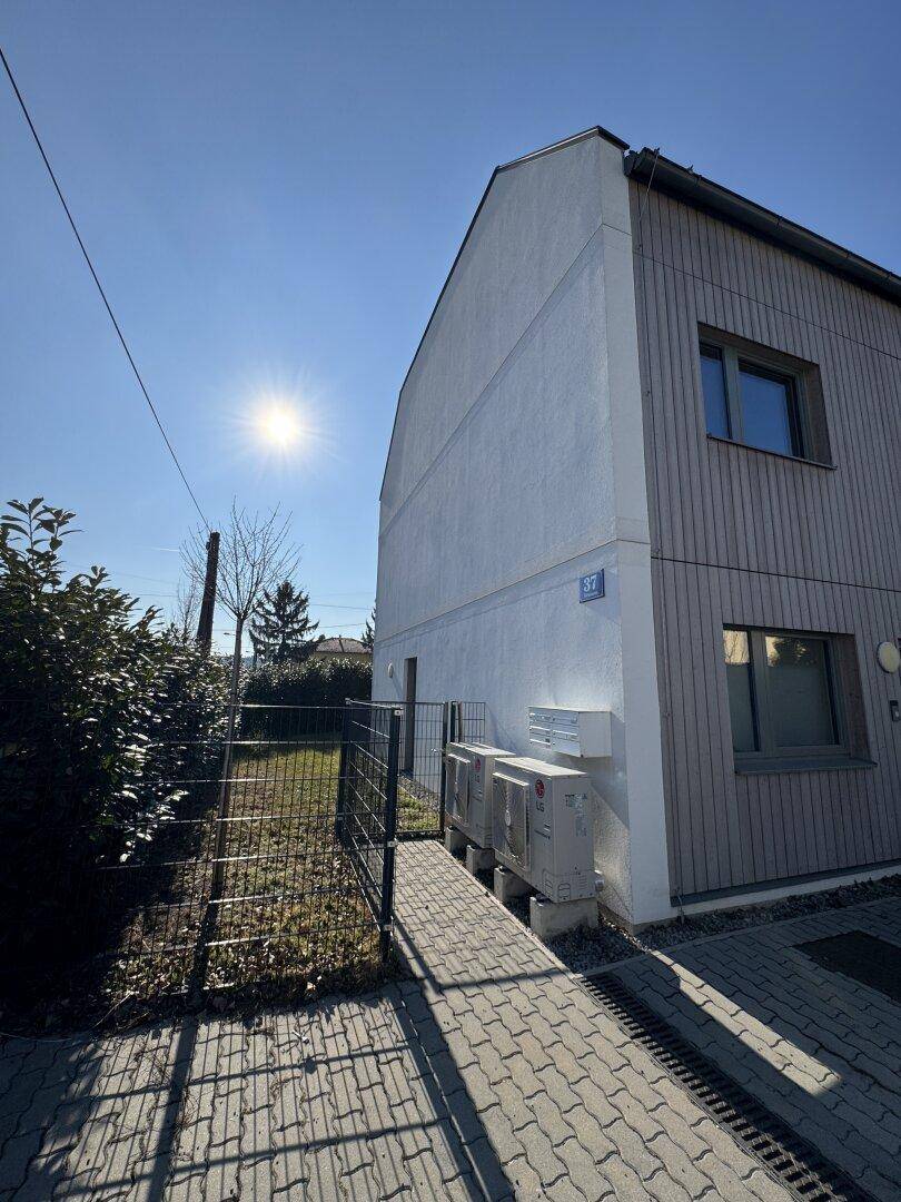 Gebäudeseite mit weißer Putzfassade und Holzverkleidung, sichtbaren Klimaanlagen und einem Zaun.
