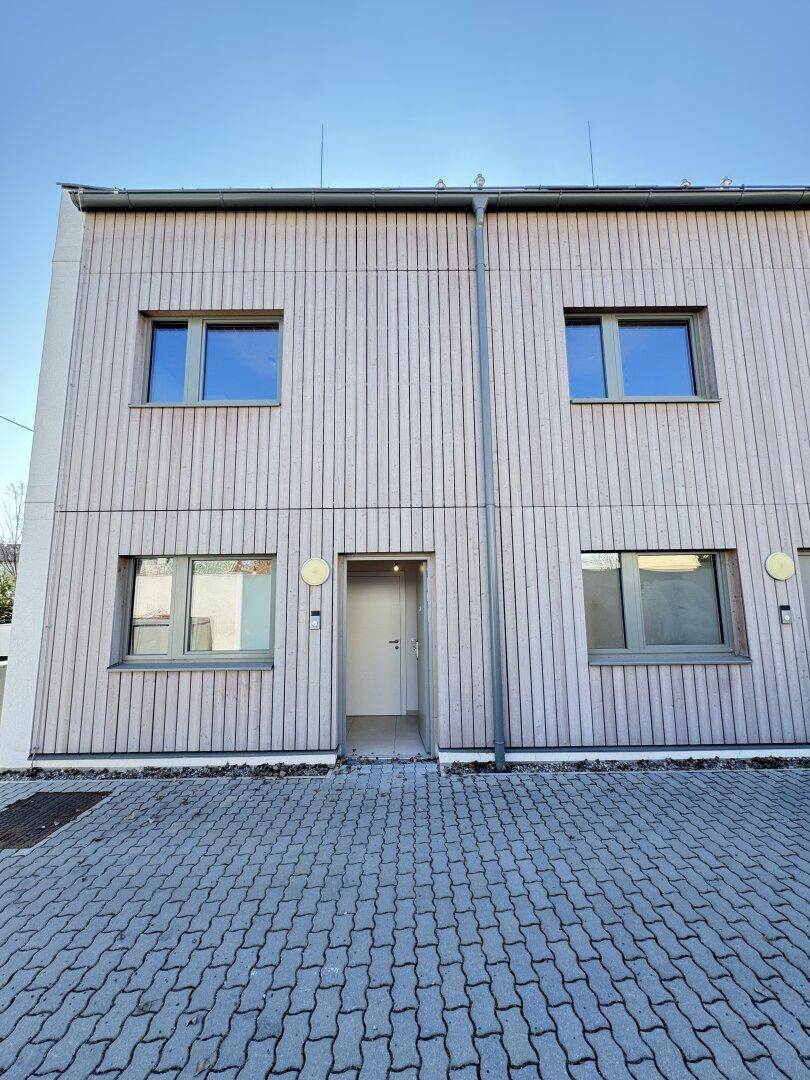 Moderne Gebäudefassade mit Holzverkleidung und mehreren Fenstern, davor ein gepflasterter Eingangsbereich.