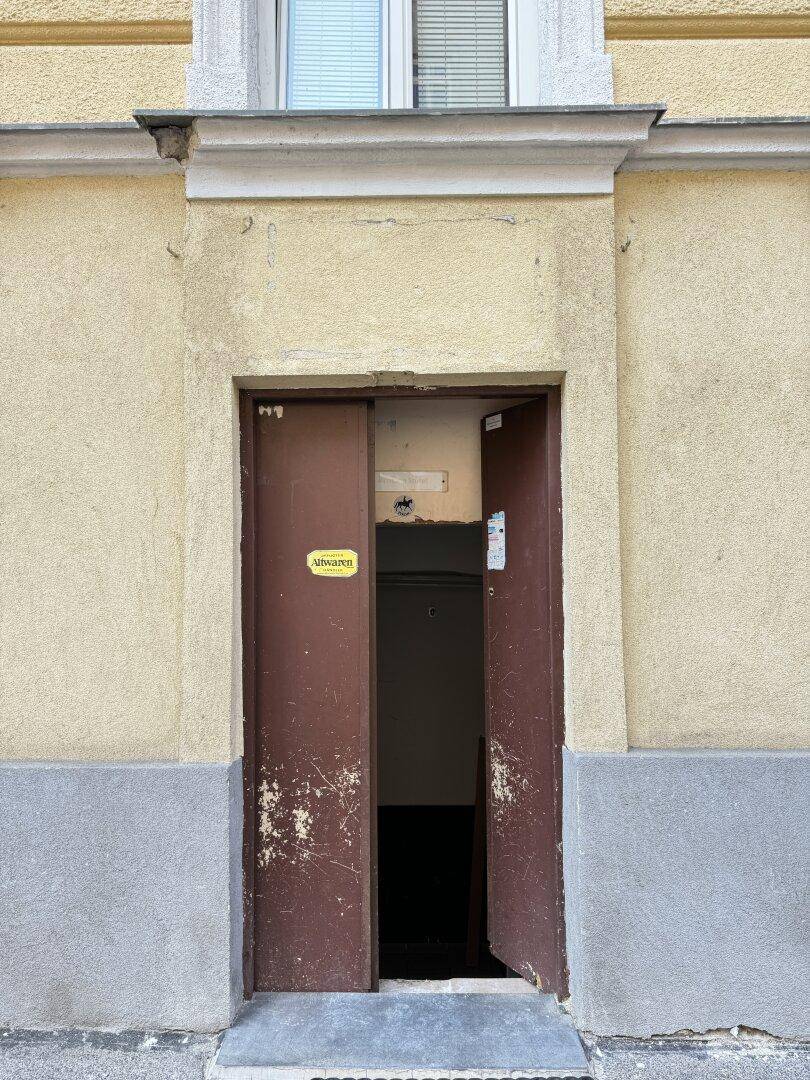 Gebäudeeingang mit einer abgenutzten braunen Tür, gelber Fassade und grauem Sockelbereich.