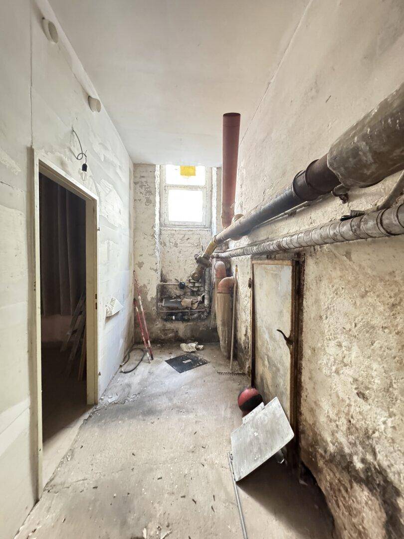 Schmaler, unfertiger Raum mit freiliegenden Rohren, einem kleinen Fenster und Betonboden, wahrscheinlich ein Kellerbereich.