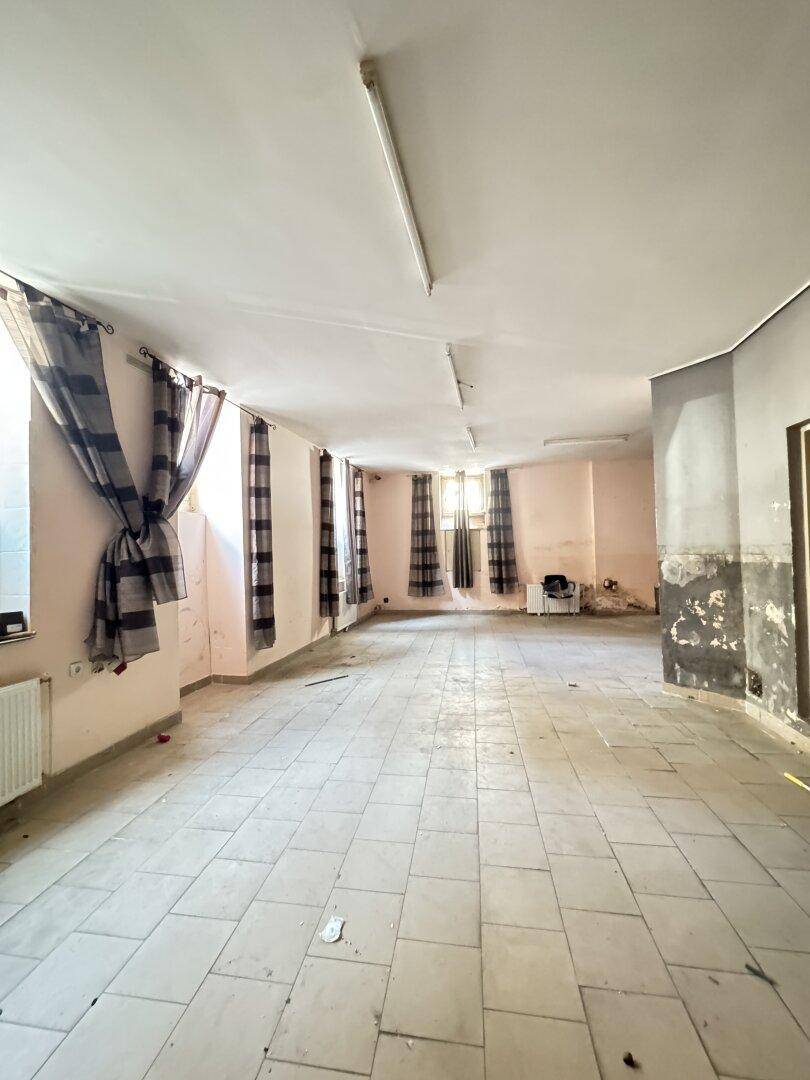 Langer, leerer Raum mit Fliesenboden, Vorhängen an den Fenstern und Deckenbeleuchtung, in renovierungsbedürftigem Zustand.