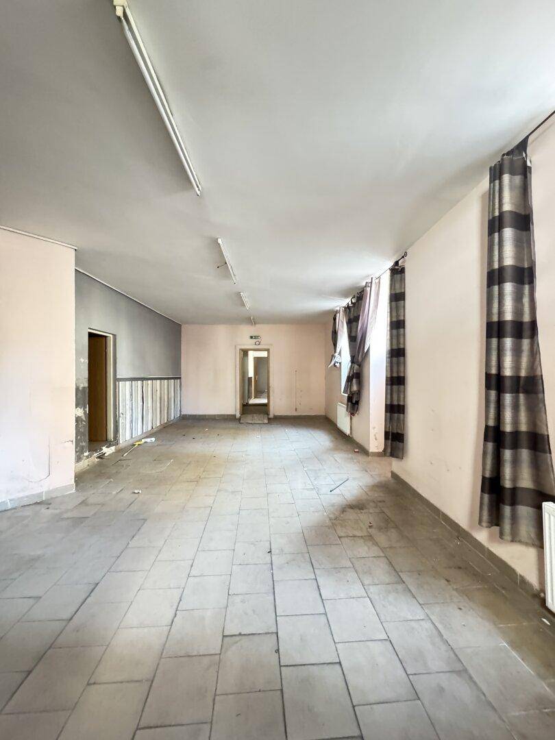 Langer, leerer Raum mit Fliesenboden, Vorhängen an den Fenstern und Deckenbeleuchtung, in renovierungsbedürftigem Zustand.