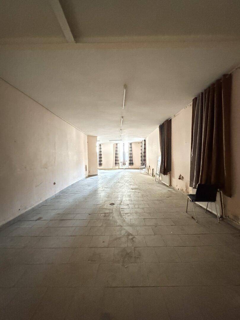 Langer, leerer Raum mit Fliesenboden, Vorhängen an den Fenstern und Deckenbeleuchtung, in renovierungsbedürftigem Zustand.