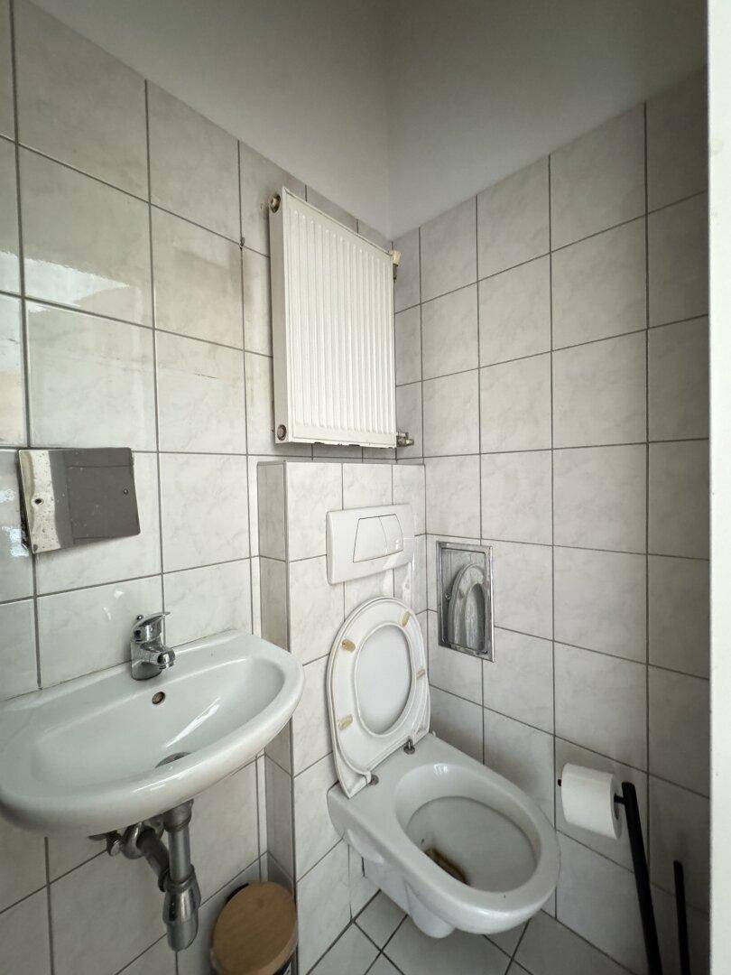 Kleines WC mit Wand-WC, Waschbecken und Heizkörper, einfach gefliest und funktional ausgestattet.