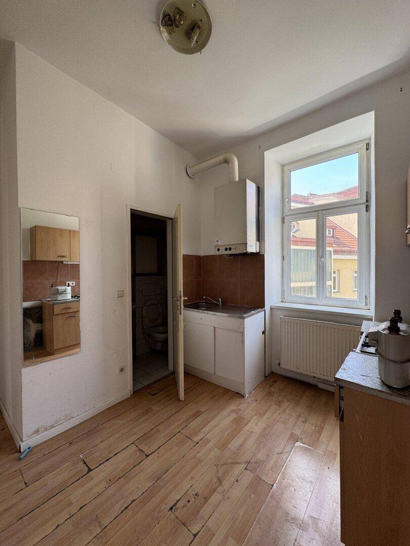 Einfache Küche mit Gastherme, Spülbecken und Fensterblick auf die umliegenden Gebäude, renovierungsbedürftig.