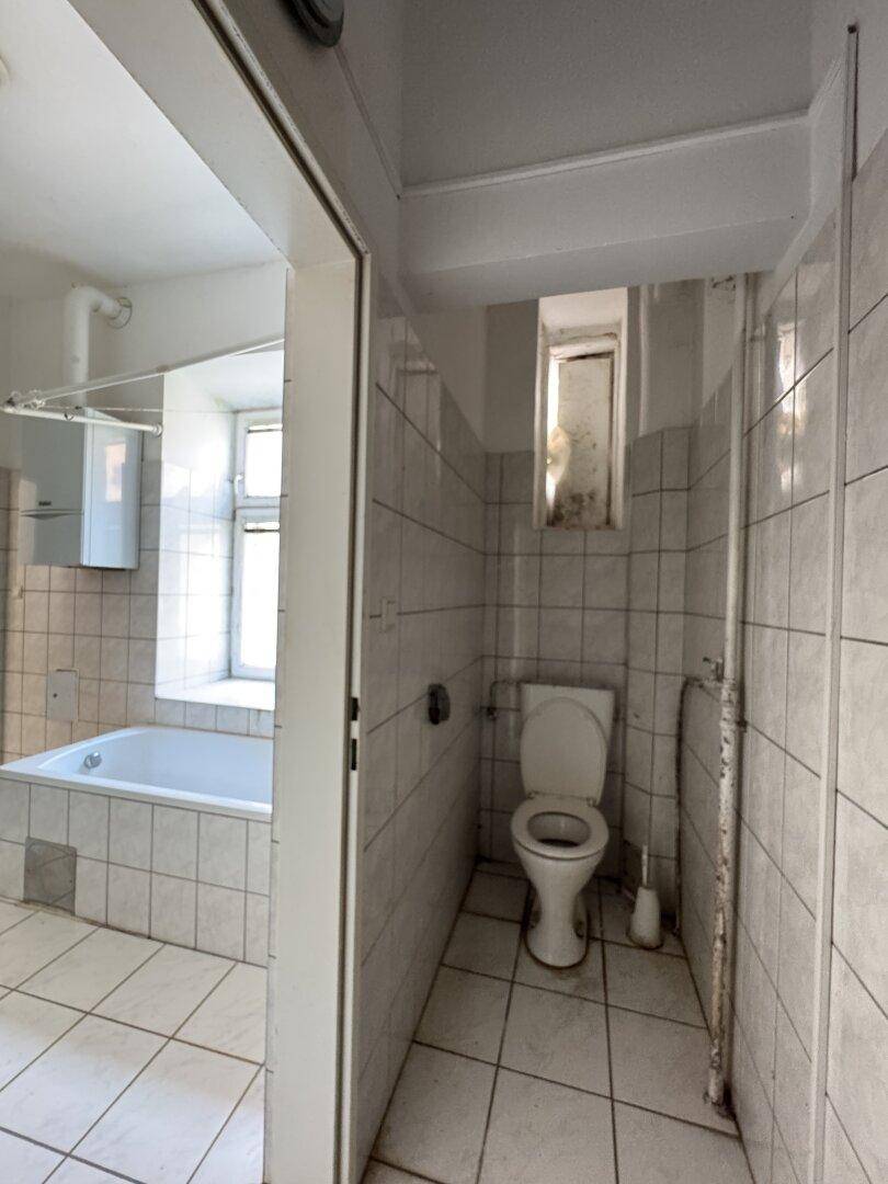 Badezimmer mit Badewanne und separatem WC-Bereich, gefliest und mit Fenster, das renovierungsbedürftig ist.