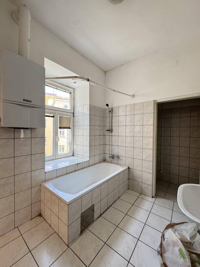 Badezimmer mit Badewanne, Fenster und Gastherme, komplett gefliest und renovierungsbedürftig.