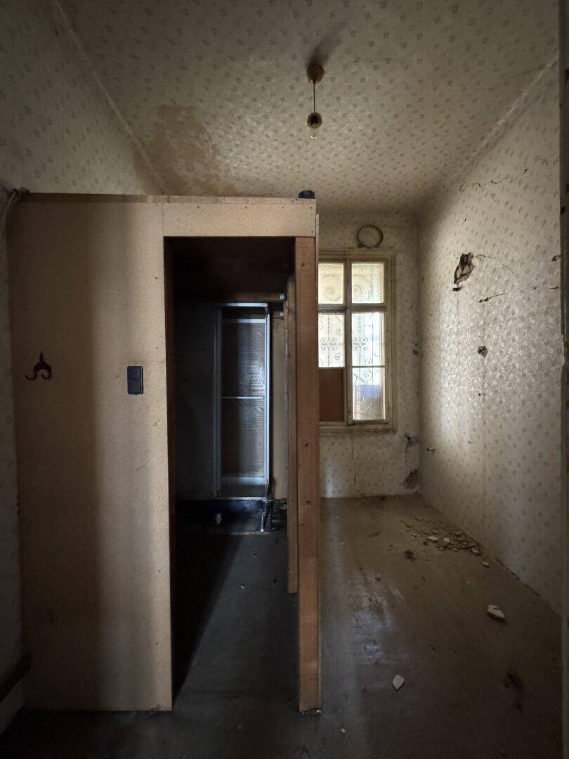 Dunkler Flur mit alter Einbaustruktur und Fenster mit Gitter, renovierungsbedürftig.