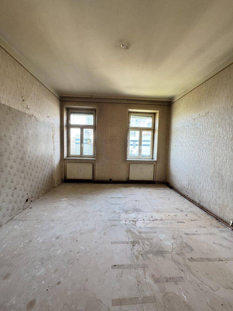 Heller, unmöblierter Raum mit zwei Fenstern und abgelöster Tapete, wartet auf Renovierung.