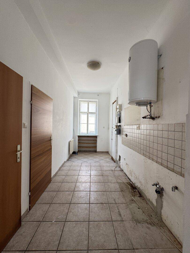 Langer Flur mit Fliesenboden, Fenster am Ende und Warmwasserboiler an der Wand.