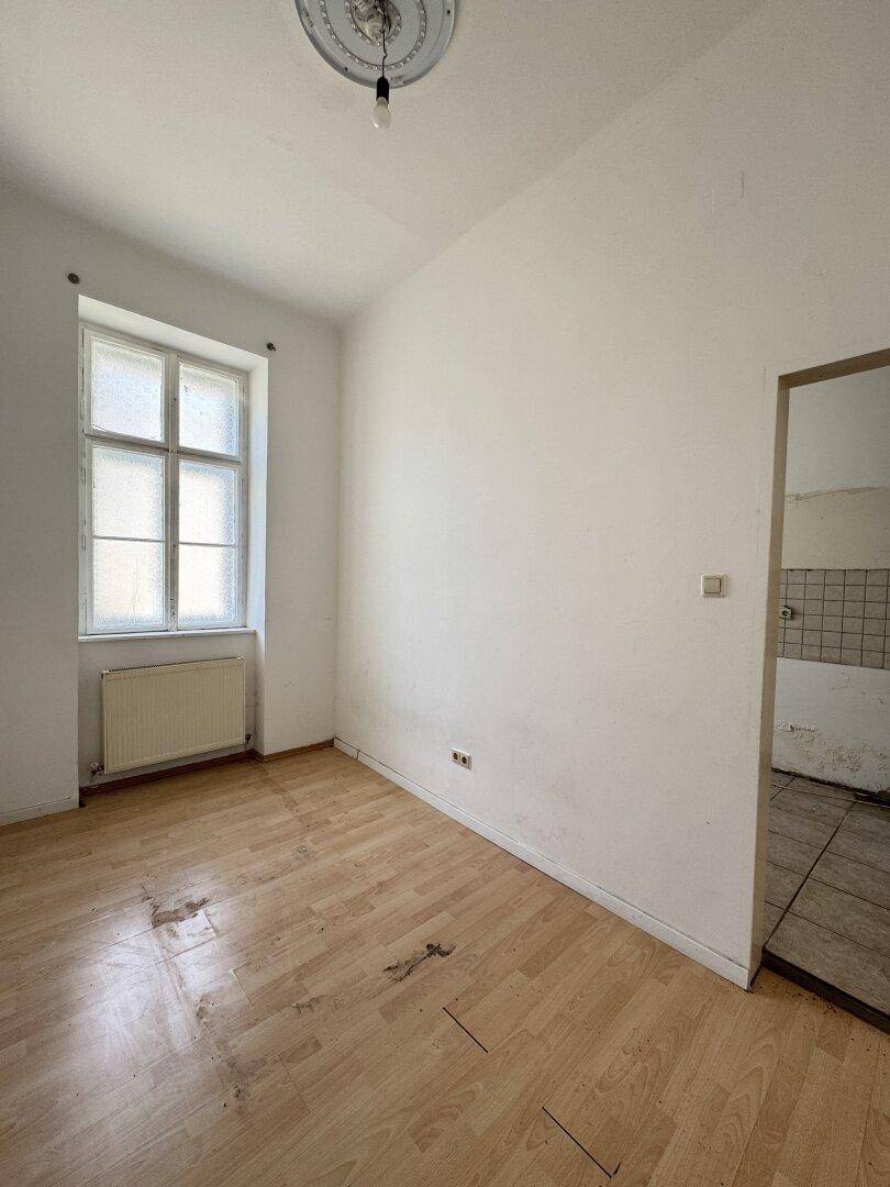 Helles Zimmer mit Holzboden und einem Fenster, angrenzend an einen gefliesten Bereich.